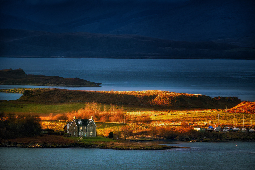 Schottisches Küstenhaus bei Oban | Ein traditionelles Steinhaus steht prominent am Ufer einer Bucht nahe Oban, Schottland. Die Szene wird von warmem, goldenem Licht beleuchtet, das die umliegenden Felder und Hügel hervorhebt, während der Hintergrund in tiefen Blautönen liegt. Boote sind am Ufer einer nahegelegenen Anlegestelle zu sehen, was die maritime Umgebung unterstreicht. - Realisiert mit Pictrs.com