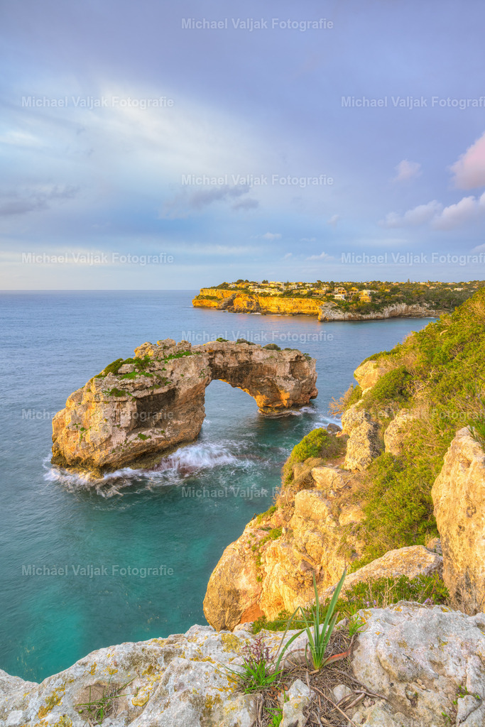 Es Pontàs auf Mallorca im Morgenlicht | Die Sonne steigt langsam über den Horizont und lässt Es Pontàs in warmen Goldtönen erstrahlen. Der Felsbogen wirkt in diesem Moment besonders kraftvoll, während das Meer darunter sanft im neuen Licht glitzert. Die Farben des Himmels mischen sich mit der Ruhe des Morgens – ein stiller Augenblick, in dem Mallorca seine natürliche Schönheit offenbart. - Realisiert mit Pictrs.com