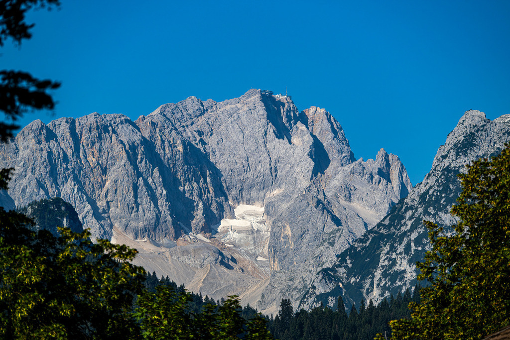 Gipfel der Erhabenheit: Deutschlands Höchster im Fokus | Eingerahmt von sattem Grün erheben sich die mächtigen, zerklüfteten Felswände der Zugspitze imposant gegen den tiefblauen Himmel. Auf dem schneidigen Gipfel zeichnen sich die Bergstation und der Sendemast deutlich ab, während weiter unten die Reste des Höllentalferners im Felskessel schimmern. Das Licht- und Schattenspiel auf dem rauen Wettersteinkalk verleiht der Aufnahme eine eindrucksvolle Tiefe. Ein Bild, das die Sehnsucht nach den Bergen, pure Wildheit und magische alpine Momente weckt. - Realisiert mit Pictrs.com
