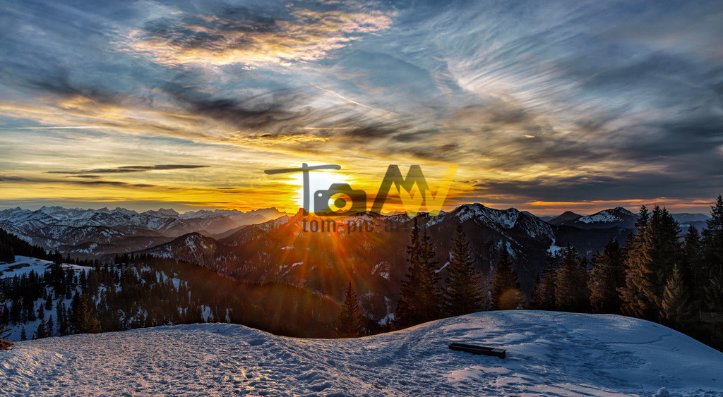 Sonnenaufgang im Winter-Bayrische Alpen--Tegernsee---Berge | Das Bild zeigt einen malerischen Sonnenuntergang in einer winterlichen Berglandschaft. Es handelt sich um eine Aufnahme, die wahrscheinlich im bayerischen Alpenraum, möglicherweise in der Nähe des Tegernsees, entstanden ist. Die Szenerie zeigt verschneite Hügel im Vordergrund und eine Bergkette im Hintergrund, beleuchtet durch die tief stehende Sonne.Die Aufnahme fängt dramatische Wolkenformationen und intensive Lichtstrahlen ein. - Realisiert mit Pictrs.com