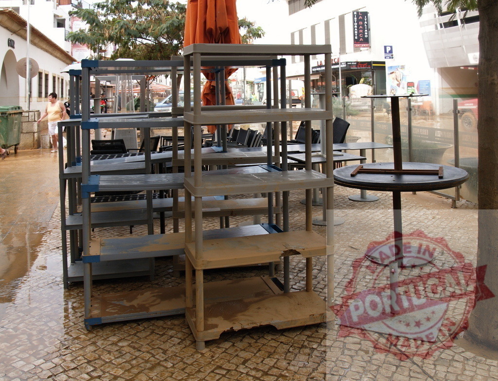 Muddy shelves and tables after flood damage | Albufeira Flood - discruction by weather problems Outdoor cafe items sit covered in mud and debris after a devastating flood - Realized with Pictrs.com