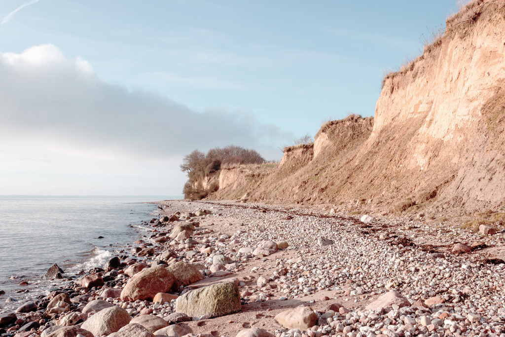 Wandbild: Steilküste in pastellartigen Farben | Dieses Wandbild im Querformat zeigt die Steilküste am Meer. Die Farben auf diesem Bild sind pastellartig und dezent. Das leicht rötliche beige der Abbruchkante bringt eine schöne wohnliche Wärme ins Wandbild. Am Naturstrand sind viele Steine zu sehen. Der Himmel ist hellblau. Direkt am Horizont ist der Himmel durch den Sonnenschein noch heller. Eine schöne weißgraue Wolke befindet sich am Himmel. Schaffen Sie ein maritimes Ambiente in Ihrem Wohnzimmer und holen Sie sich dieses stilvolle Wandbild. Es Wandbild ist auf Leinwand, Aluminium-Platte, Acrylglas oder als Holzdruck erhältlich. Die Wandbilder werden individuell für Sie in vielen Abmessungen produziert. Daher passen die Ostseekult Wandbilder immer perfekt an Ihre Wände. - Realisiert mit Pictrs.com
