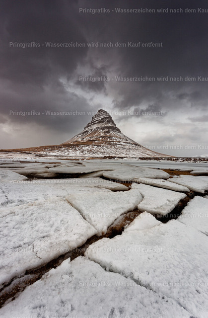 Kirkjufell unter dramatischem Himmel | Der markante Berg Kirkjufell erhebt sich aus einer winterlichen Landschaft in Island. Schneeflächen, dunkle Wasserläufe und der schwere Himmel verstärken die raue, kraftvolle und fast mystische Stimmung dieses bekannten isländischen Naturmotivs. - Realisiert mit Pictrs.com