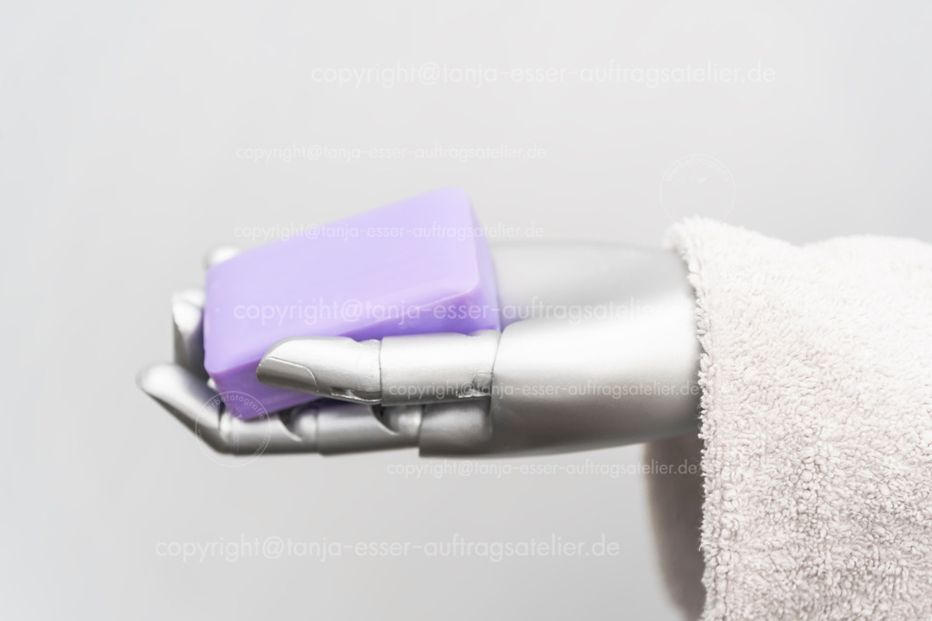 Robot takes care of people | Der Roboterarm hält Frotteehandtücher und ein Stück Seife in der Hand. Das Bild symbolisiert die Pflege von Menschen durch Maschinen. Konzeptuelles Foto vor weißem Hintergrund mit Copy Space.
