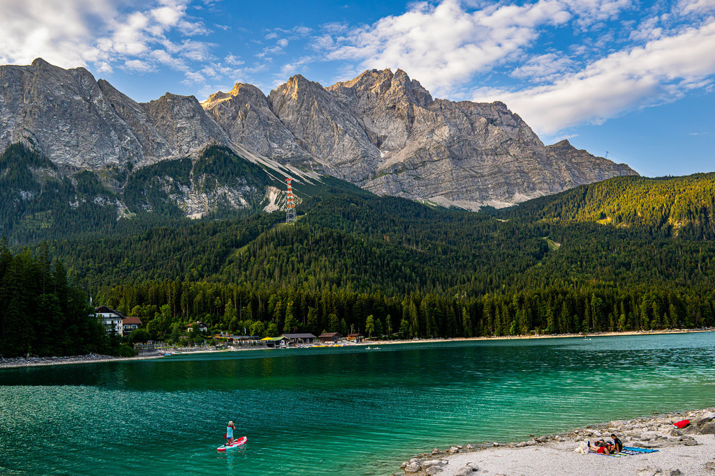 Alpenidylle pur: Sommerglück am Eibsee | Ein strahlender Sommertag entfaltet sich am malerischen Eibsee am Fuße des mächtigen Zugspitzmassivs. Das smaragdgrüne Wasser lädt zu erfrischenden Aktivitäten wie Stand-Up-Paddling ein, während die Sonne die markanten Felsgipfel in goldenes Licht taucht. Saftig grüne Tannenwälder säumen das Ufer und kleine, idyllische Hütten versprechen Ruhe und Entspannung. Dieses Foto fängt die pure Lebensfreude, die Sehnsucht nach Bergen und See sowie die unberührte Schönheit der Natur ein. - Realisiert mit Pictrs.com