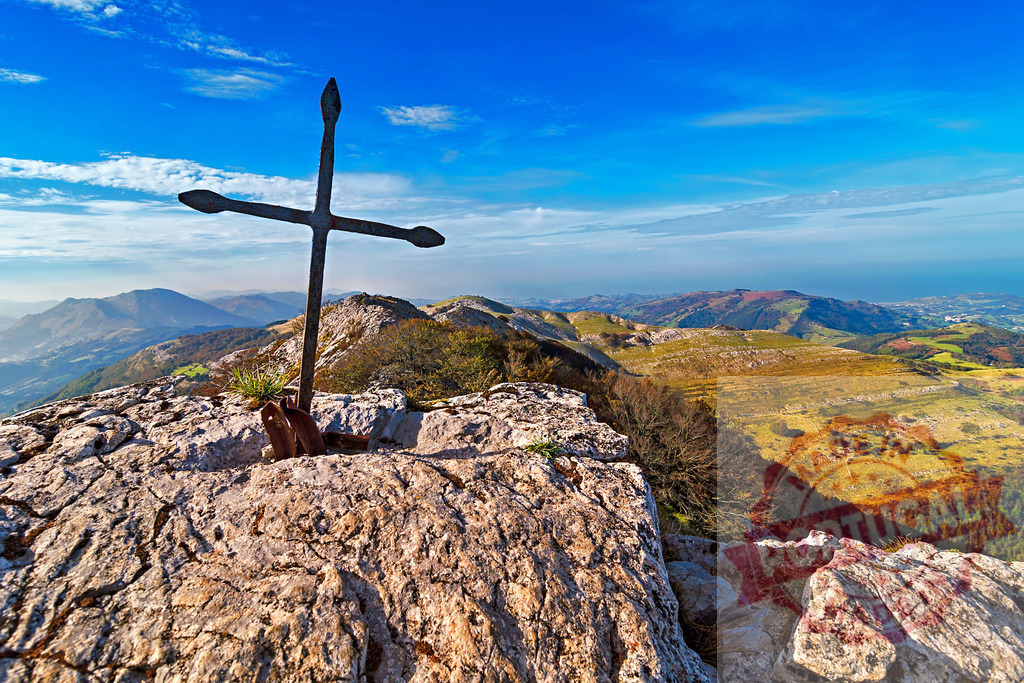 Large metal cross standing on a rocky mountain peak overlooking a vast landscape | Scenic view of a metal cross on a mountain peak, symbolizing faith and achievement against a backdrop of rolling hills and blue sky - Realisiert mit Pictrs.com
