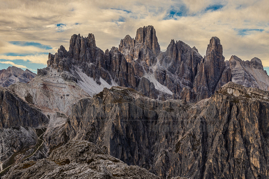 R5M29203_20251004 | Dieses beeindruckende Querformat zeigt die majestätische und raue Bergwelt der Dolomiten, vermutlich die Cadini di Misurina Gruppe, in einer Übergangszeit zwischen Herbst und Winter. Die Szenerie wird dominiert von einer Kette schroffer, gezackter Felsgipfel, die sich dramatisch gegen einen bewölkten Himmel abzeichnen. Vereinzelte Schneefelder haften an den steilen Flanken und in den Mulden, was auf die fortgeschrittene Jahreszeit hindeutet. Im Vordergrund erstreckt sich eine karge, felsige Landschaft mit Geröll und spärlicher Vegetation, durchzogen von einem gewundenen Pfad, der sich die Hänge hinaufschlängelt. Die warmen Erdtöne der Felsen kontrastieren mit dem kühlen Grau des Himmels und dem Weiß des Schnees und verleihen dem Bild eine kraftvolle, ursprüngliche Atmosphäre. - Realisiert mit Pictrs.com