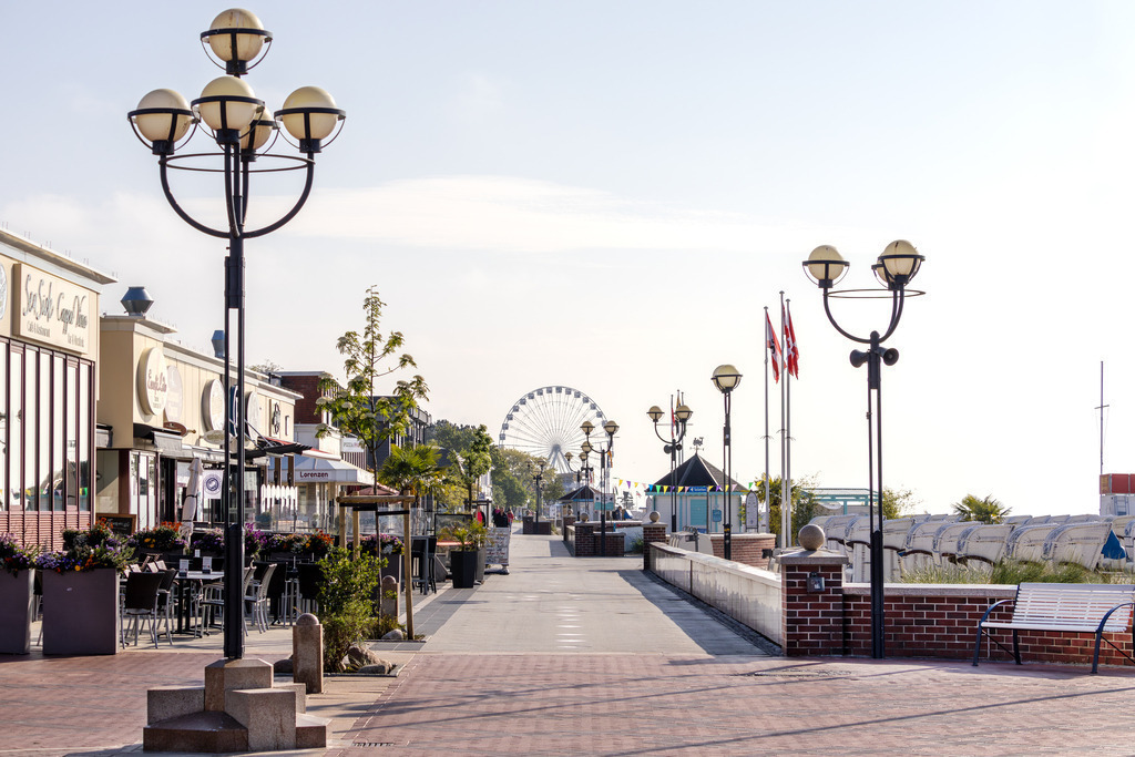 Wandbild: Grömitzer Morgenstimmung – Promenade erwacht | Dieses maritime Wandbild zeigt die Promenade von Grömitz am frühen Morgen – noch ruhig, bevor der Tag beginnt. Links reihen sich Cafés und Geschäfte mit einladenden Außenbereichen, während rechts einige Strandkörbe aufgereiht stehen und auf ihre ersten Gäste warten. Moderne Straßenlaternen, gepflegte Pflanzkübel und wehende Fahnen verleihen der Szene ein frisches, lebendiges Flair. Im Hintergrund erhebt sich ein großes Riesenrad, das dem Bild eine verspielte, touristische Note verleiht. Die klare Luft und das sanfte Licht des Morgens schaffen eine entspannte Atmosphäre, die zum Flanieren und Verweilen einlädt. Dieses Motiv bringt die Leichtigkeit eines Ostseeurlaubs direkt an die Wand und eignet sich ideal für maritime Wohnkonzepte. Erhältlich als Leinwandbild, Acrylglasbild, Alu-Dibond FineArt Print oder als Akustikbild – ein stilvoller Blickfang für Zuhause, Büro oder Ferienwohnung. - Realisiert mit Pictrs.com