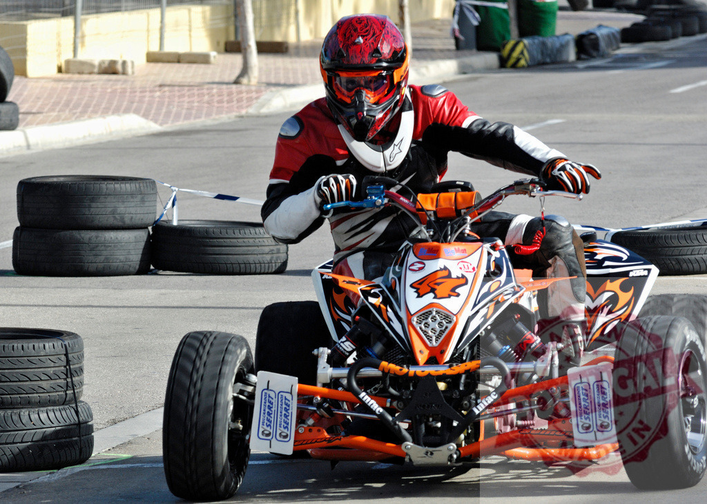 Pilot driving quad bike on asphalt track near tires | Quad bike pilot competing in a race on an asphalt track, wearing protective gear and focused on driving - Realisiert mit Pictrs.com