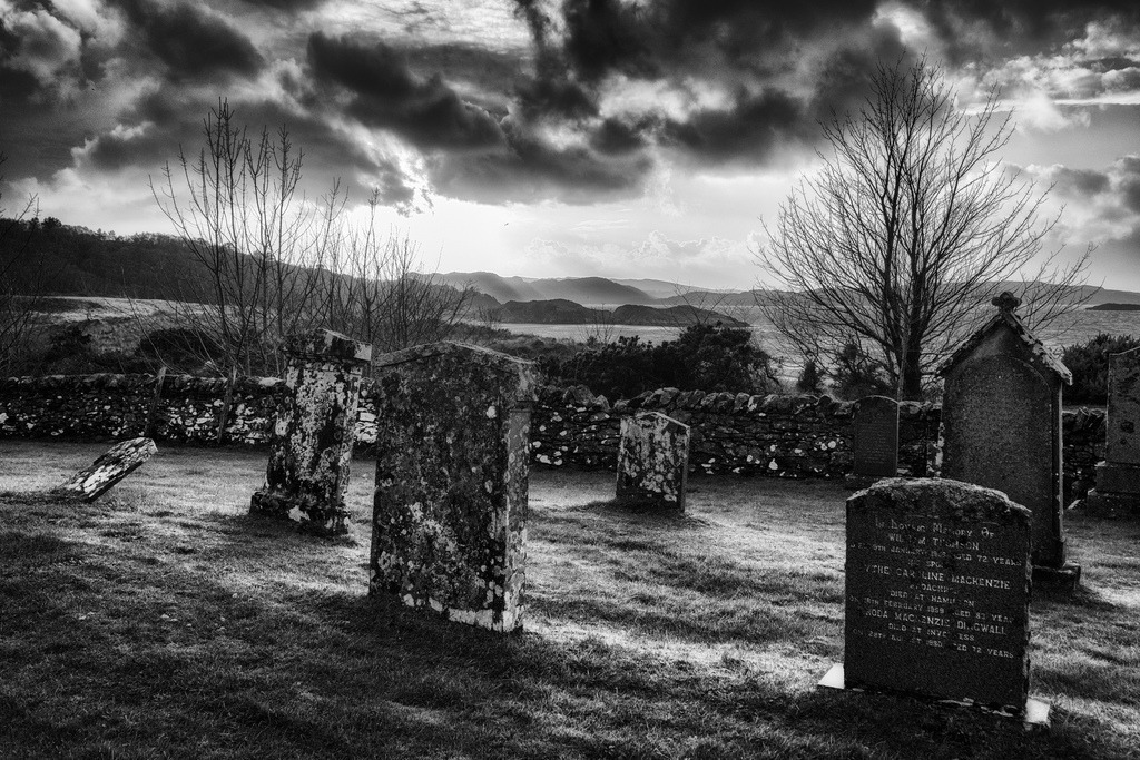 Friedhof Gairloch | Eine Reihe alter, moosbewachsener Grabsteine steht auf einem grasbewachsenen Friedhof in Gairloch, Schottland. Im Hintergrund erstreckt sich eine Steinmauer, kahle Baeume und eine weite Landschaft mit einem Gewaesser und huegeligen Ufern unter einem dramatischen, bewoelkten Himmel. Die Schwarz-Weiss-Darstellung betont die Texturen und die melancholische Stimmung der Szenerie. - Realisiert mit Pictrs.com