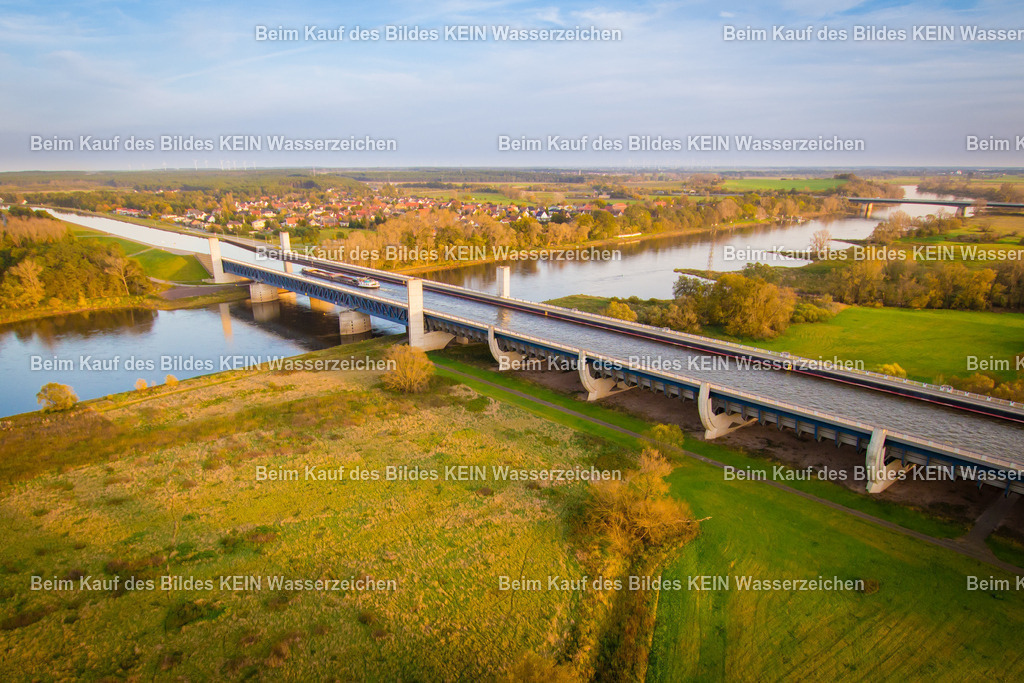 Trogbrücke-5843 | Das Luftbild zeigt die Trogbrücke des Wasserstraßenkreuzes Magdeburg aus einer Drohnenperspektive. Das Bauwerk wird vom warmen orangefarbenen Licht geprägt und hebt sich eindrucksvoll von der umgebenden Landschaft ab. Aus der Vogelperspektive werden die klaren Linien der Brückenkonstruktion und die außergewöhnlichen Dimensionen der Wasserstraße besonders deutlich sichtbar.Die Trogbrücke wurde 2003 eröffnet und ist mit einer Gesamtlänge von 918 Metern die längste Kanalbrücke Europas. Der wassergefüllte Brückentrog führt den Mittellandkanal über die Elbe und ermöglicht der Binnenschifffahrt eine durchgehende Verbindung zwischen den west- und ostdeutschen Wasserstraßen. Der Trog ist 34 Meter breit und rund 4,25 Meter tief, sodass auch große Gütermotorschiffe und Schubverbände die Brücke sicher passieren können. Für den Bau wurden rund 24.000 Tonnen Stahl und etwa 68.000 Kubikmeter Beton verwendet.Die Drohnenaufnahme verdeutlicht eindrucksvoll die Dimensionen und die technische Leistungsfähigkeit dieses außergewöhnlichen Ingenieurbauwerks. Die Kombination aus moderner Stahlbaukunst, innovativer Wasserbautechnik und dem warmen Licht macht die Trogbrücke zu einem der bedeutendsten technischen Wahrzeichen des Wasserstraßenkreuzes Magdeburg. - Realisiert mit Pictrs.com