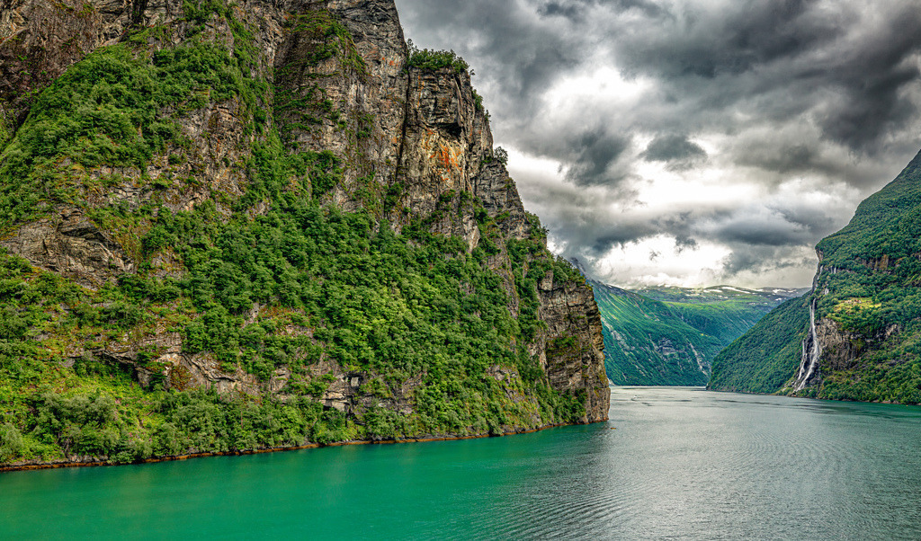 Passage im Geiranger Fjord,blaues Wasser,grüne Felsen,Norwegen | Das Bild zeigt den Geirangerfjord in Norwegen. Dieser Fjord ist bekannt für seine beeindruckende Naturkulisse und gehört seit 2005 zum UNESCO-Weltnaturerbe. Der Fjord ist etwa 15 Kilometer lang und von bis zu 800 Meter hohen, steilen Felswänden umgeben, die direkt ins Wasser abfallen. Er wurde während mehrerer Eiszeiten durch Gletscher geformt, die tiefe Täler in die Landschaft schnitten.  - Realisiert mit Pictrs.com