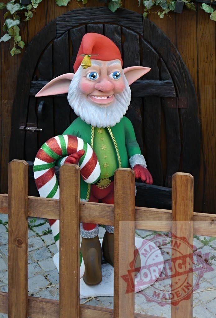Christmas elf holding candy cane standing behind wooden fence | Christmas elf with red hat and green clothes holding big candy cane standing behind wooden fence in front of wooden door - Realized with Pictrs.com