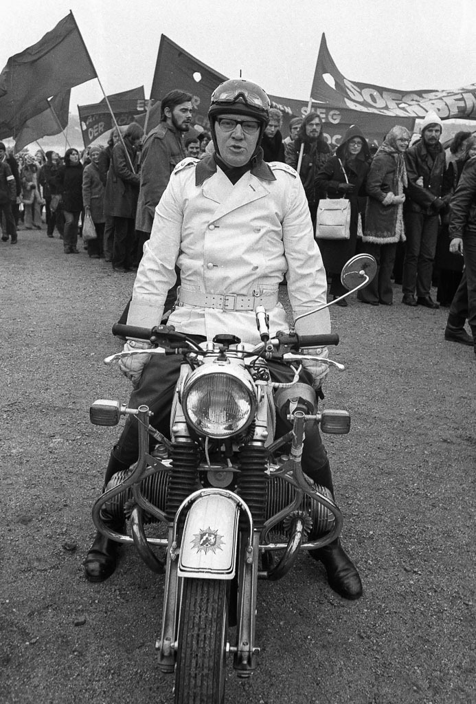 013-1973-007 | 25.000 Menschen demonstrierten am 14.1.1973 in Bonn gegen den Vietnamkrieg. Dazu aufgerufen hatten Gruppen aus dem linksradikalen Spektrum der sogenannten K-Gruppen. Der Protest gegen den Vietnamkrieg und die Kriegsführung der USA bildete seit den späten 1960er Jahren eine entscheidende Triebkraft aller außerparlamentarischen Aktionen in Westdeutschland und Westberlin. Das Foto zeigt einen Polizisten auf seinem Motorrad, während sich Demonstrationsteilnehmer im Hintergrund sammeln. - Realisiert mit Pictrs.com