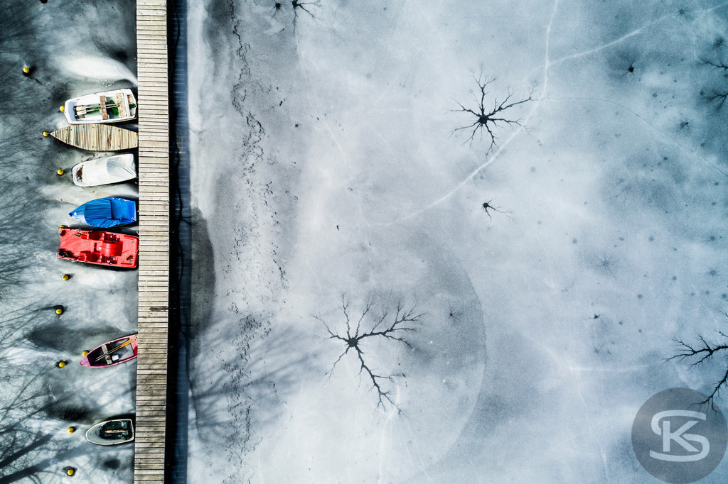 Eingefrorene Boote am Wintersteg – Drohnenaufnahme eines zugefrorenen Sees | Beeindruckende Luftaufnahme von eingefrorenen Booten am Steg eines zugefrorenen Sees. Die Drohnenfotografie zeigt winterliche Stille mit Baumschatten auf der Eisfläche aus der Vogelperspektive – atmosphärische Winterimpression. - Realisiert mit Pictrs.com