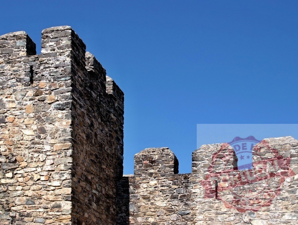 Medieval castle stone wall with battlements under clear blue sky. Region Norte - Portugal | Ancient stone fortress wall with crenelated battlements standing against a vibrant blue sky - Realized with Pictrs.com