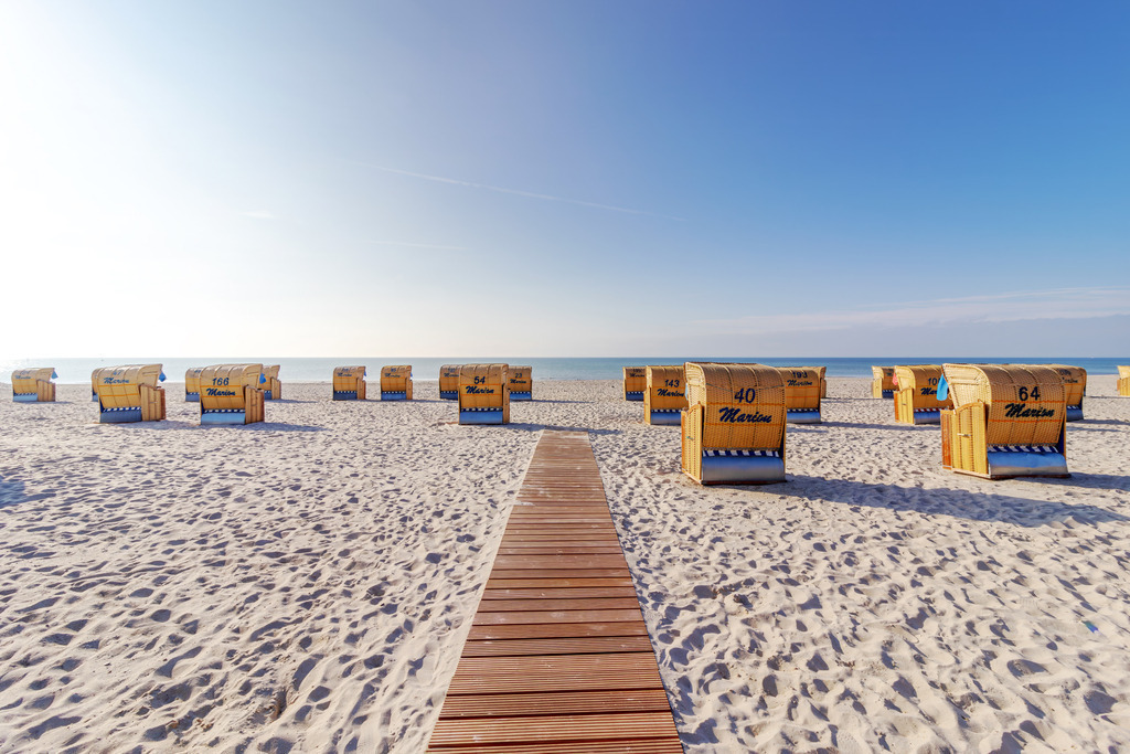 Leinwandbild Urlaubsfreunde – Strandkörbe am Meer in Grömitz | Dieses maritime Wandbild zeigt den Strand von Grömitz an einem sonnigen Tag. Ein hölzerner Steg führt geradlinig durch den Sand Richtung Meer und gliedert die Szene in klare Perspektiven. Links und rechts stehen zahlreiche Strandkörbe in ordentlichen Reihen und verleihen dem Bild eine strukturierte, ruhige Atmosphäre. Der Sand ist hell und fein, das Meer liegt ruhig, und der Himmel zeigt sich mit zarten Schleierwolken freundlich und weit. Die Komposition vermittelt Gelassenheit und Urlaubsstimmung – ein typisches Bild der Ostseeküste. Dieses Motiv eignet sich ideal als Wandbild für maritime Wohnkonzepte und bringt die entspannte Atmosphäre eines Sommertages direkt ins Zuhause.  - Realisiert mit Pictrs.com