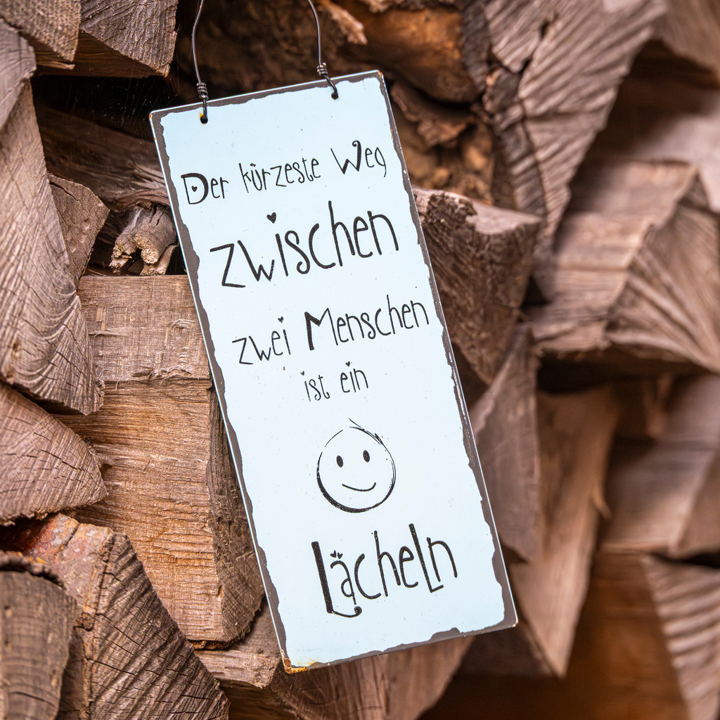 Lächeln | Vor einem rustikalen Hintergrund aus grob gestapeltem, verwittertem Brennholz hängt ein schmales Blechschild in hellem Pastellblau. Die handgeschriebene Botschaft „Der kürzeste Weg zwischen zwei Menschen ist ein Lächeln“ strahlt eine herzliche Wärme aus, die einen sanften Kontrast zur harten, trockenen Textur des Holzes bildet. Ein kleines, gezeichnetes Smiley unterstreicht die freundliche Aura dieses ländlichen Stilllebens. - Realisiert mit Pictrs.com