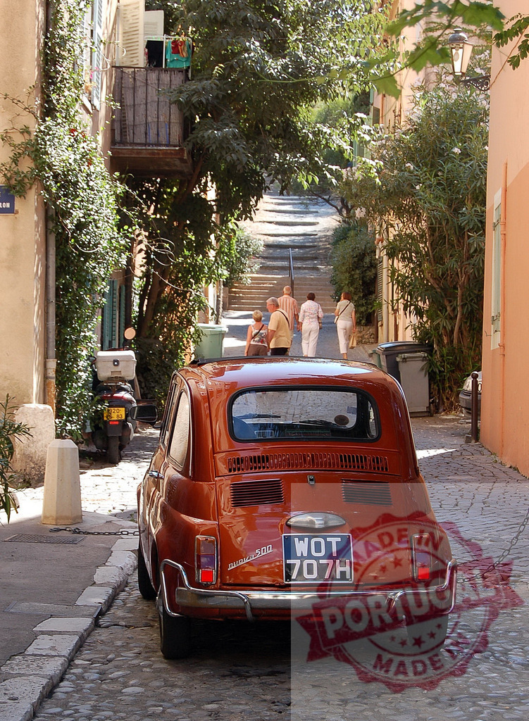 Classic Fiat 500 parked in narrow cobblestone street in France with tourists walking uphill | Orange vintage Fiat 500 parked on a narrow street with cobblestones and tourists walking uphill in a typical French cityscape - Realized with Pictrs.com
