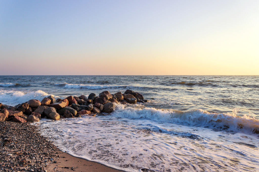 Wandbild: Sonnenaufgang an der Ostsee | Dieses Wandbild im Querformat zeigt einen schönen Sonnenaufgang an der Ostsee. An der Steinmole bricht sich eine Welle. Der Himmel leuchtet in einem schonen warmen rotorange.  - Realisiert mit Pictrs.com