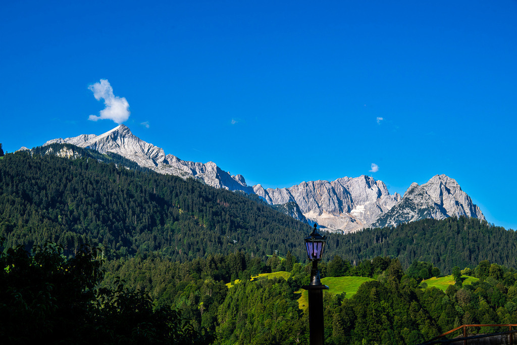 Majestätische Gipfelparade: Von der Alpspitze zur Zugspitze | Unter einem makellos blauen Sommerhimmel entfaltet sich die volle Pracht des Wettersteingebirges. Auf der linken Seite zieht die charakteristische Pyramide der Alpspitze die Blicke auf sich, gekrönt von einer kleinen, eleganten Wolke. Der Blick wandert weiter über den kühnen Grat hin zu den mächtigen Felsmassiven der Zugspitze und der Waxensteine. Im Vordergrund schaffen saftig grüne Bergwälder, sanfte Almen und eine klassische Lataniere einen stimmungsvollen Rahmen. Eine Szenerie, die zeitlose Ruhe, alpine Erhabenheit und bayerische Idylle perfekt vereint. - Realisiert mit Pictrs.com