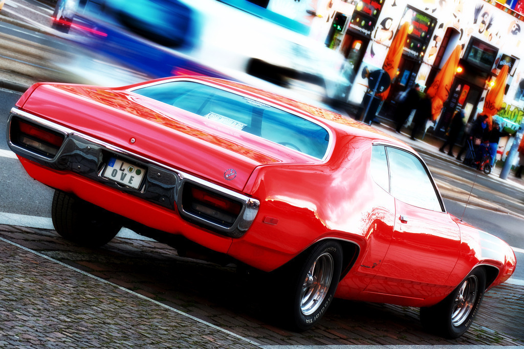 Red Oldie | Der rote Oldtimer im Vordergrund reflektiert das grelle Tageslicht während der im Hintergrund vorbeifahrende Polizeiwagen und die Menschen vor der bunten Häuserfassade vom dynamischen Treiben in der Großstadt zeugen.Unsere Empfehlung: Leinwand, Alu-Dibond oder Acrylglas - Realisiert mit Pictrs.com