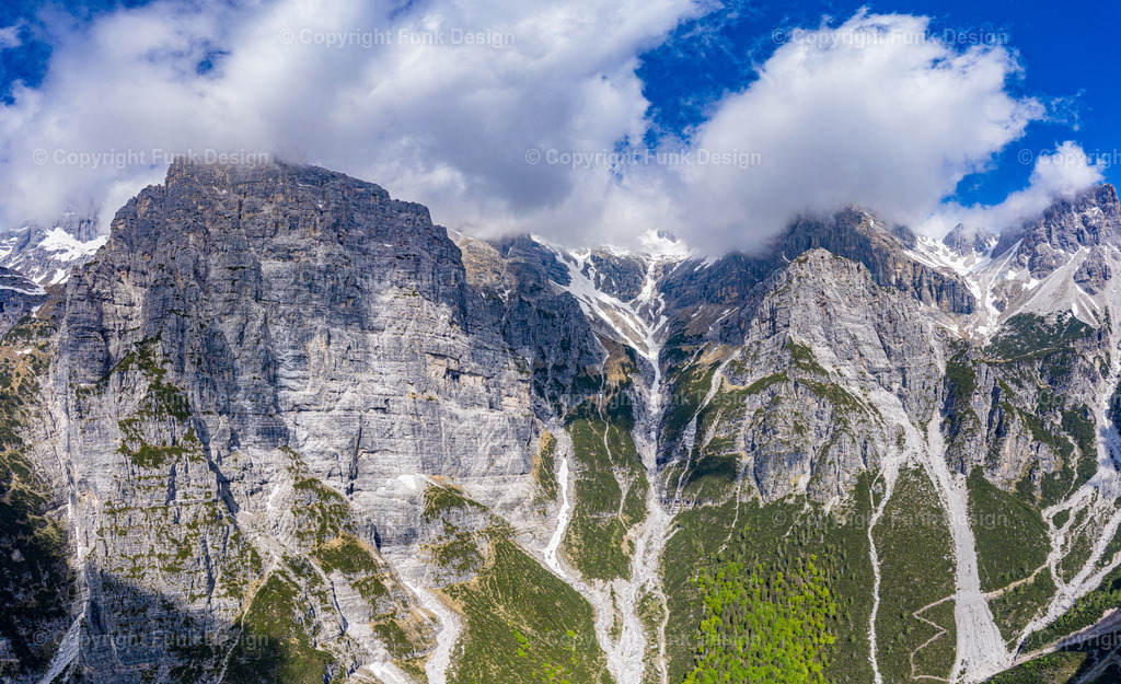 Wolken über schroffen Gipfeln – Alpen, Italien | Massive Felswände ragen steil in den Himmel, während dichte Wolken die Gipfel teilweise verschlucken. Schneereste und Geröllrinnen zeichnen die Berge wie Linien nach und geben dem Motiv eine raue, majestätische Stimmung.
