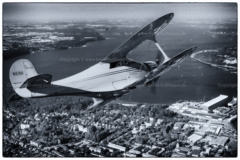 PLB_air2air_118_1827modnk2_75x50 | Luftbild. Historischer Doppeldecker Typ Beechcraft Staggerwing über Flensburg. Im Bildvordergrund - die Flensburger Nordstadt mit der Werft ( FSG ) ___ Bildanmutung - Duoton blau, farbreduziert. - Realisiert mit Pictrs.com