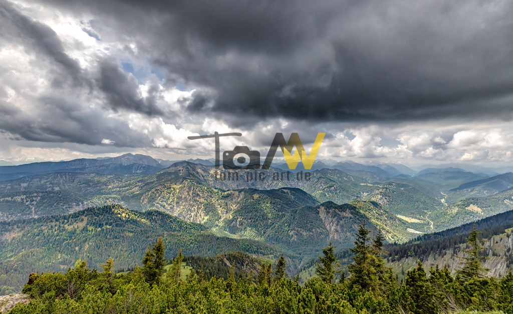 Panoramablick über das Alpenvorland |Tegernsee | Panoramablick über die Gipfel der Berge in das Alpenvorland hinab des Tegernsee.Die Aufnahme zeigt eine weite Berglandschaft mit üppiger Vegetation und dramatischen Wolken am Himmel. Die Region Tegernsee ist bekannt für ihre malerischen Ausblicke und zahlreiche Wandermöglichkeiten.  - Realisiert mit Pictrs.com