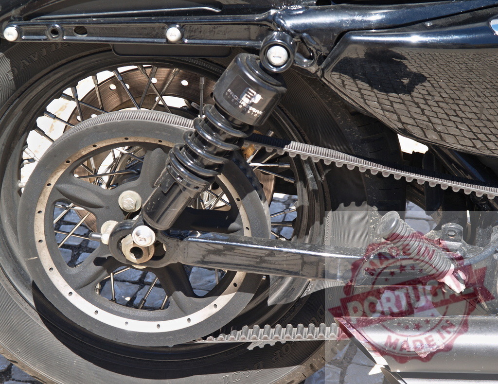Black motorcycle wheel with shock absorber and drive belt | Motorcycle rear wheel showing suspension, drive belt, and exhaust pipe detail - Realized with Pictrs.com