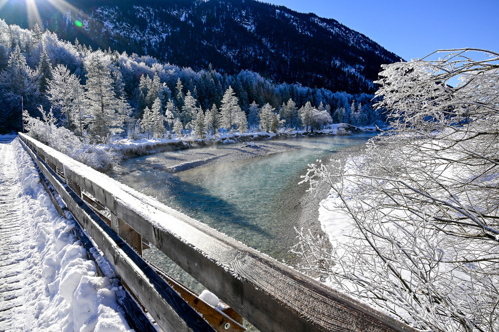 Brücke ins Winterwunderland: Magie aus Eis und Licht | Ein magischer Wintermoment, der die Zeit stillstehen lässt: Von einer tief verschneiten Holzbrücke schweift der Blick über die kristallklare, türkisblaue Isar, von deren Oberfläche sanfte Nebelschleier aufsteigen. Am Ufer im winterlichen Isartal glitzert dichter Raureif auf den Bäumen wie Millionen winziger Diamanten, während die strahlende Wintersonne ein wärmendes Licht auf die eisige Märchenlandschaft wirft. Dieses Bild fängt die vollkommene, eiskalte Ruhe und die unberührte Poesie eines perfekten Wintertages in den Alpen ein. - Realisiert mit Pictrs.com