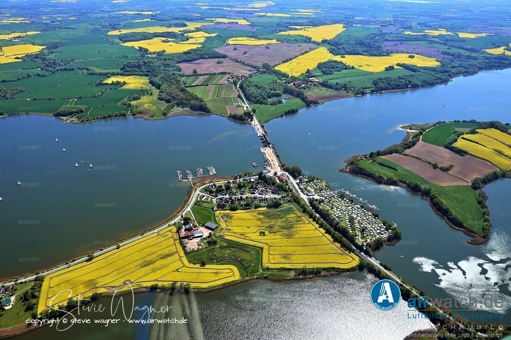 Luftbild Ostseefjord Schlei, Gunnebyer Noor, Badestrand Gunneby, Landschaftsschutzgebiet Schwansener Schleilandschaft | Die Lindaunisbrücke, auch bekannt als Schleibrücke Lindaunis, ist eine kombinierte Eisenbahn- und Straßenklappbrücke, die die Halbinseln Schwansen und Angeln an der engsten Stelle der Schlei verbindet. Sie liegt im Gemeindeteil Lindaunis der Gemeinde Boren in Schleswig-Holstein und wird von der Bahnstrecke Kiel–Flensburg und der Landesstraße L 283 genutzt. Die Brücke ist 120 Meter lang und besteht aus einem Stahlfachwerk- und einem Klappbrückenteil, wobei die Klappbrücke für den Schiffsverkehr geöffnet wird.