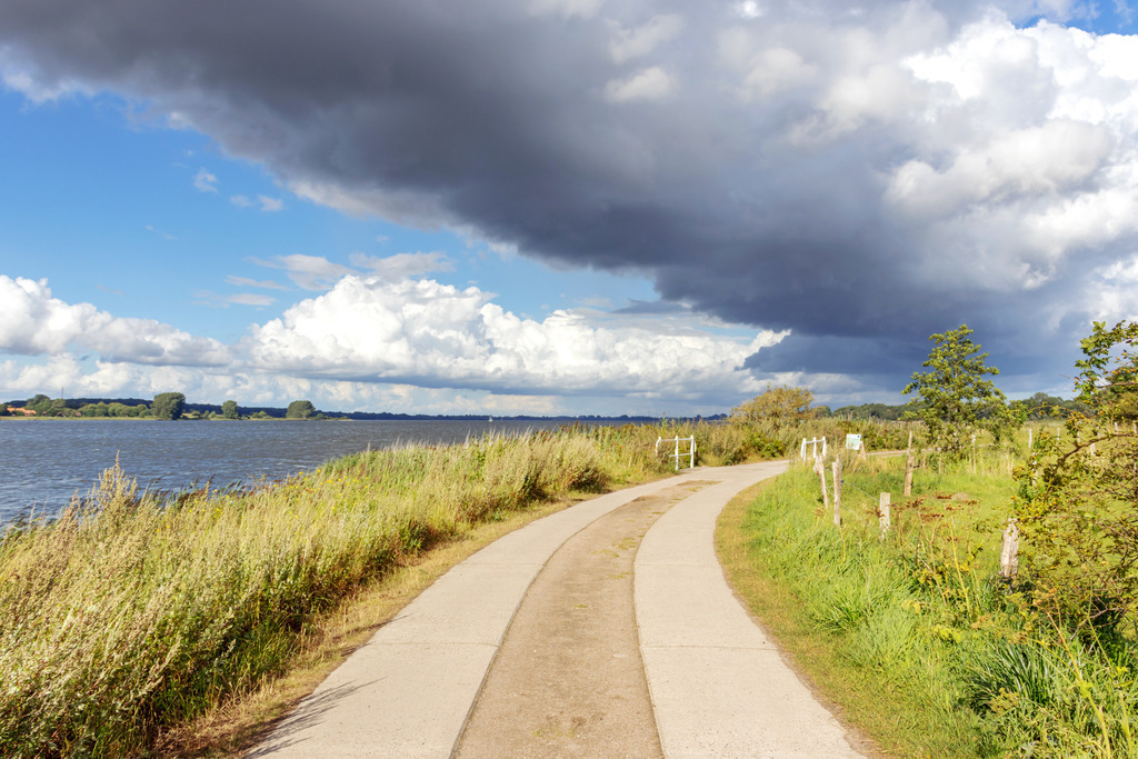 XXL Wandbild: Sonne und Wolken am Ostseefjord | Dieses XXL Wandbild im Querformat zeigt einen schönen Spazierweg am Ostseefjord. In der Mitte des Wandbilds ist ein schöner Plattenweg zu sehen. Am Himmel befindet sich eine große Wolke in unterschiedlichen Grautönen. Die Schlei befindet sich auf der linken Seite - Realisiert mit Pictrs.com