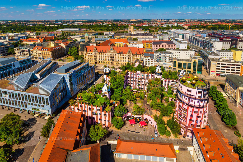 Hundertwasserhaus Grüne Zitadelle Nord-LB Bank und Souvenir Domplat Magdeburg-3474 | Die Magdeburger Innenstadt aus der Vogelperspektive: Im Mittelpunkt dieser sommerlichen Luftaufnahme steht die Grüne Zitadelle am Breiten Weg. Das farbenfrohe Hundertwasserhaus hebt sich mit seinen geschwungenen Formen, den unterschiedlichen Fassadenfarben und den begrünten Dachbereichen deutlich von den umliegenden Gebäuden ab. Links und dahinter sind moderne Geschäfts- und Bürohäuser sowie historische Gebäude mit ihren markanten Dachlandschaften zu erkennen. –  – Der Breite Weg zählt zu den bekanntesten Straßen Magdeburgs. Er durchquert die Innenstadt und verbindet bedeutende Bereiche des Stadtzentrums. Die Aufnahme zeigt, wie unterschiedlich sich die Bebauung präsentiert: historische Fassaden mit roten Ziegeldächern stehen neben modernen Geschäftshäusern und großflächigen Gebäudekomplexen. Von oben wird besonders deutlich, wie eng diese verschiedenen Bauformen im Zentrum zusammenliegen. –  –  Die Grüne Zitadelle wurde nach Entwürfen des österreichischen Künstlers Friedensreich Hundertwasser gestaltet und 2005 eröffnet. Sie war eines seiner letzten fertiggestellten Architekturprojekte. Ihren Namen verdankt sie unter anderem den begrünten Dachflächen und ihrer charakteristischen Gestaltung, die sich bewusst von der geradlinigen Architektur der Umgebung abhebt. –  – Diese Luftaufnahme vermittelt einen Überblick über einen Teil der Magdeburger City. Sie zeigt nicht nur ein bekanntes Gebäude, sondern auch dessen städtebauliche Umgebung mit Geschäftsvierteln, Straßen und unterschiedlichen Architekturepochen. So lässt sich die Innenstadt aus einer Perspektive betrachten, die vom Straßenraum aus kaum möglich ist. - Realisiert mit Pictrs.com
