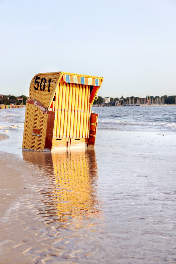 Wandbild: Strandkorb mit Spiegelung in Eckernförde | Dieses Wandbild im Hochformat zeigt einen Strandkorb am Sandstrand in Eckernförde, der durch leichtes Hochwasser teilweise im Meer stehen. Durch das schöne morgendliche Sonnenlicht ergibt sich eine  Spiegelung des Strandkorbs im flachen Wasser.  - Realisiert mit Pictrs.com