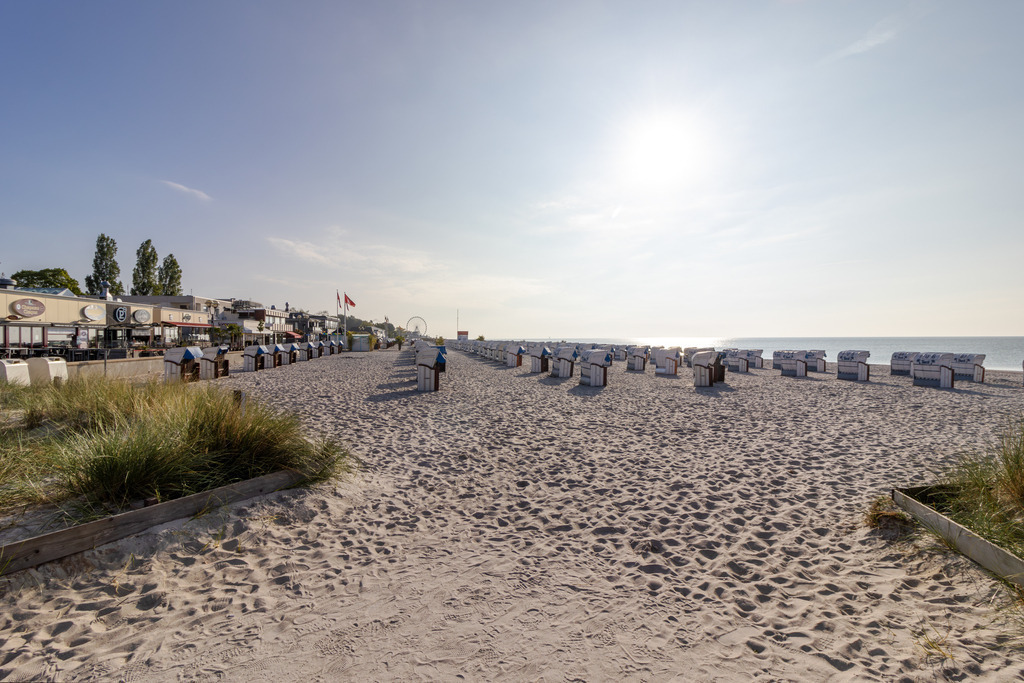 Wandbild: Zwischen Gräsern und Horizont – Grömitz ganz still | Dieses Wandbild zeigt eine ruhige Strandszene in Grömitz, eingefangen in den frühen Morgenstunden. Im Vordergrund erstreckt sich ein sandiger, leicht bewachsener Küstenbereich mit hohem Gras, der den Blick auf den Strand sanft rahmt. Zahlreiche Strandkörbe stehen in geordneten Reihen und richten sich zum Meer aus – bereit für einen Tag voller Ruhe und Erholung. Links im Bild befinden sich Gebäude und Strandbetriebe mit Außenterrassen und wehenden Fahnen, die dem Bild eine lebendige, aber zurückhaltende Note verleihen. Das Meer liegt ruhig im Hintergrund, der Himmel ist klar und das morgendliche Licht fällt weich über die gesamte Szene. Die Komposition lebt vom Wechselspiel aus Struktur und Weite, aus Licht und Küstenleben – ein stiller Moment der Morgenstimmung zwischen Natur und Architektur. Dieses Motiv eignet sich ideal als Wandbild für maritime Wohnkonzepte – ob als Leinwandbild, Acrylglasbild, Alu-Dibond FineArt Print oder als Akustikbild. Ein stilvoller Akzent für Wohnzimmer, Büro oder Ferienwohnung. - Realisiert mit Pictrs.com