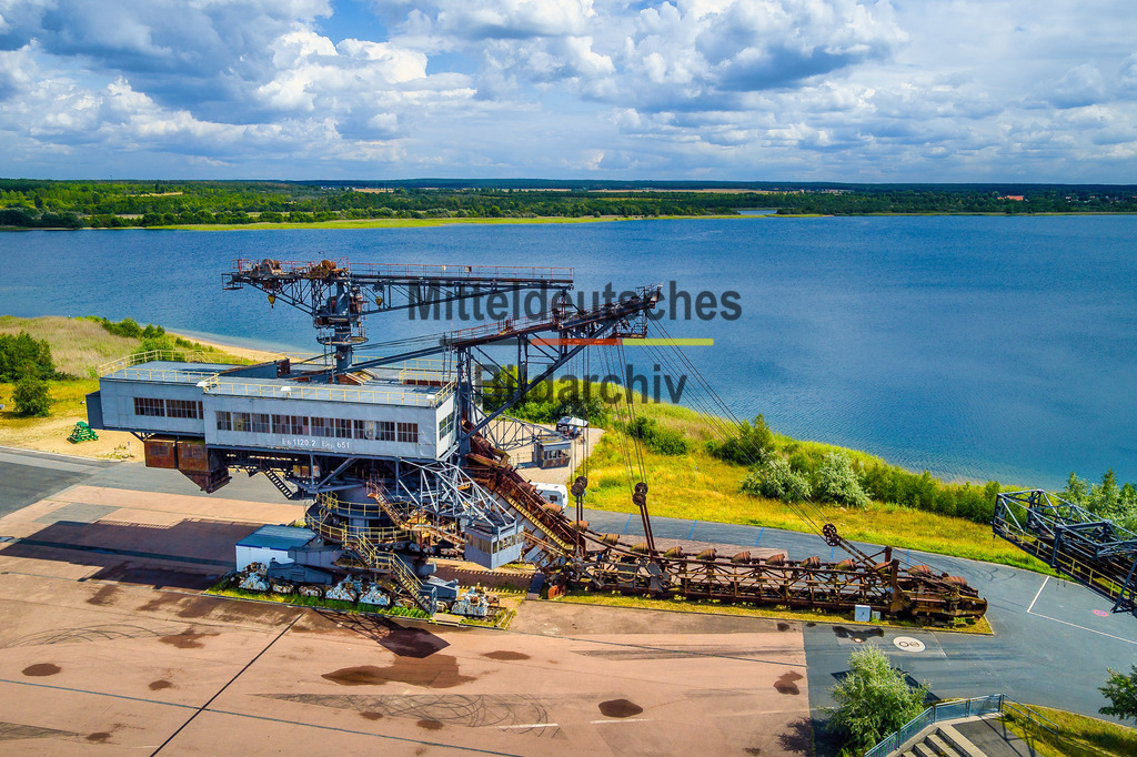 Ferropolis_Bagger_Mad_Max_Stadt_aus_Eisen-0131_GEN | Dieses Luftbild zeigt den Eimerkettenbagger „Mad Max“ in Ferropolis bei Gräfenhainichen in Sachsen-Anhalt aus einer eindrucksvollen Drohnenperspektive. Die gewaltige Maschine gehört zu den bekanntesten Exponaten der „Stadt aus Eisen“ und erinnert an die jahrzehntelange Braunkohleförderung im mitteldeutschen Revier.Mit einer Länge von rund 148 Metern und einer Höhe von etwa 52 Metern zählt „Mad Max“ zu den größten erhaltenen Tagebaugroßgeräten Europas. Charakteristisch ist die mächtige Eimerkette mit ihren zahlreichen Schaufeln, die einst ununterbrochen Erdschichten und Braunkohle bewegte. Die gewaltigen Stahlkonstruktionen vermitteln noch heute einen Eindruck von den technischen Leistungen des Bergbaus im 20. Jahrhundert.Aus der Vogelperspektive werden die Dimensionen des Baggers besonders eindrucksvoll sichtbar. Die filigran wirkenden Stahlgerüste stehen im spannenden Kontrast zu ihrer tatsächlichen Größe. Gemeinsam mit den weiteren Großgeräten von Ferropolis bildet „Mad Max“ ein einzigartiges Ensemble der Industriekultur in Sachsen-Anhalt.Dieses Luftbild zeigt eines der bekanntesten technischen Denkmäler Deutschlands und dokumentiert eindrucksvoll die Bergbaugeschichte sowie den Strukturwandel der Region rund um Gräfenhainichen und Ferropolis. - Realisiert mit Pictrs.com