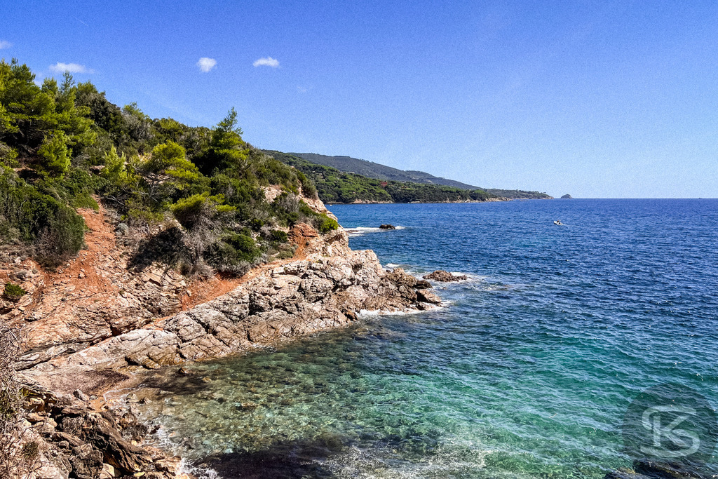 Kristallklare Buchten: Wilde Felsküste auf Elba – Wandbild | Blick auf eine zerklüftete Felsküste auf der Insel Elba; im Vordergrund kristallklares, türkisfarbenes Wasser, das in tiefes Blau übergeht, gesäumt von grüner mediterraner Macchia unter strahlend blauem Himmel. - Realisiert mit Pictrs.com