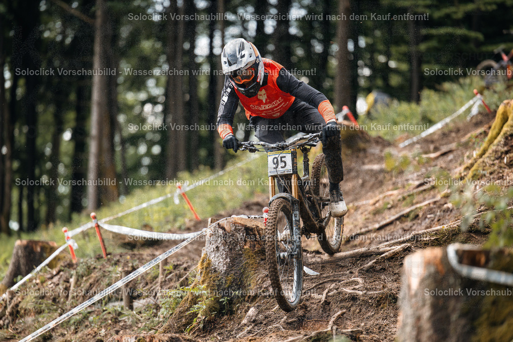 Mountainbiker im Schnee | Ein Mountainbiker in voller Ausrüstung rast einen steilen, verschneiten Hang hinunter. Er trägt einen roten Overall mit dem Schriftzug 'Race Tech' und eine schwarze Helm mit einer gelben Visier. Er trägt auch Handschuhe und Stiefel. Der Biker ist in der Mitte des Bildes zu sehen und fährt auf einem schmalen Pfad, der von Bäumen und Büschen gesäumt ist. Der Hintergrund ist verschneit und bewaldet. Der Biker scheint sich in guter Form zu befinden und genießt die Fahrt. - Realisiert mit Pictrs.com
