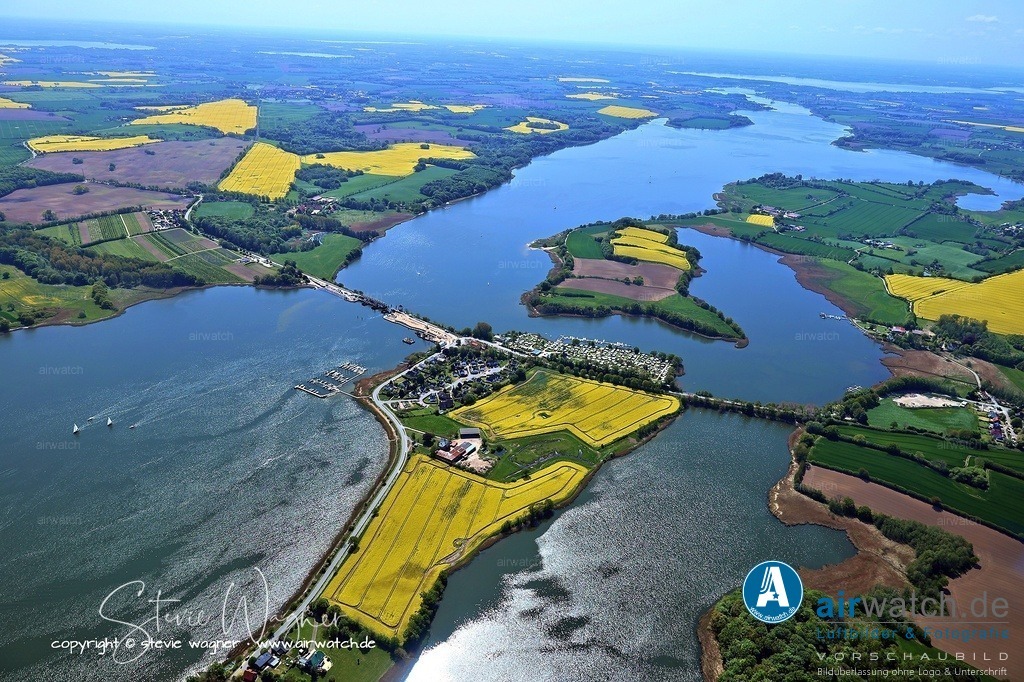 Luftbild Ostseefjord Schlei, Gunnebyer Noor, Badestrand Gunneby, Landschaftsschutzgebiet Schwansener Schleilandschaft, Schleibrücke Lindaunis | Die Geschichte von Lindau am Ufer der Schlei ist eng mit der Herrschaft Dallacker verbunden, die einst zur Herrschaft Lindau gehörte. Dort befinden sich heute noch Findlinge in der Schlei, die nach einer Sage von einem Besitzer des Hofes aufgestellt wurden, dessen elf Söhne nach Britannien ausgewandert waren, um ihren Heimkehrern und Vorüberfahrenden die Existenz des Hofes der elf Söhne zu dokumentieren. Diese Sage wird durch die Namensähnlichkeit mit Orten wie Gunby oder Great Gonerby in England unterstützt, was auf eine mögliche historische Grundlage hindeutet.