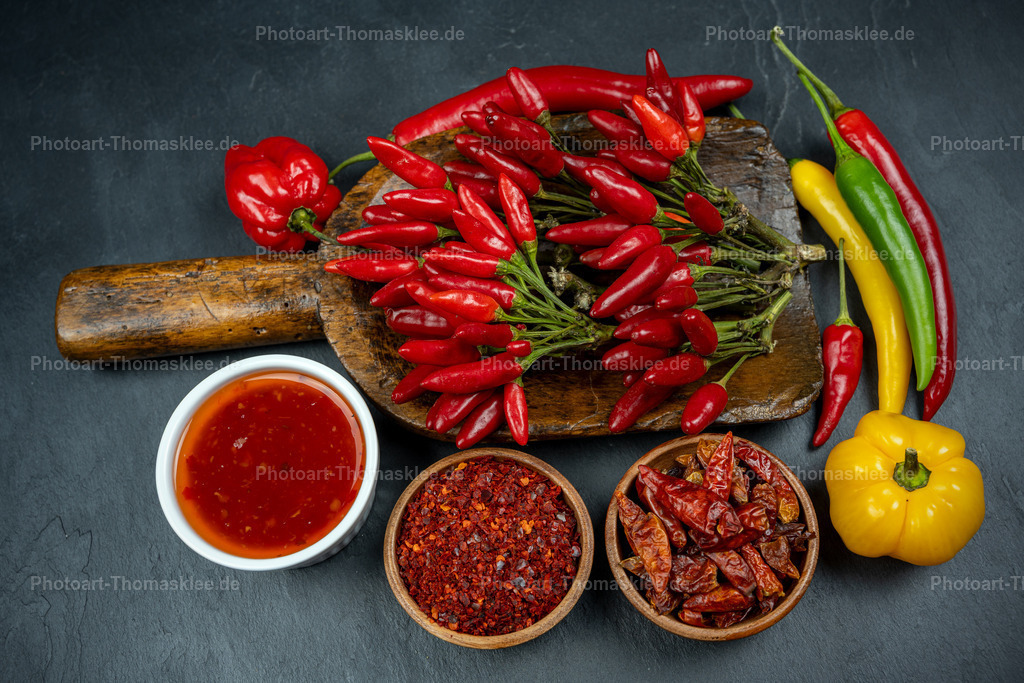 Scharfe Gewürze: Chilischoten mit Sauce und Pulver | Eine sehr alte Mehlschaufeln präsentiert einen bunten Strauß roter Chilischoten auf dunklem Schieferhintergrund. Frische gelbe und grüne Paprika ergänzen die feurige Komposition harmonisch. Eine weiße Schale enthält leuchtend rote Chilisauce, während zwei Holzschalen gemahlenes Chilipulver und getrocknete Chilistücke zeigen. Die warmen Rot- und Gelbtöne kontrastieren wirkungsvoll mit dem kühlen Schiefer. Diese kunstvolle Anordnung verschiedener Gewürze inspiriert Hobbyköche zu kreativen Kochexperimenten mit scharfen Aromen. - Realisiert mit Pictrs.com