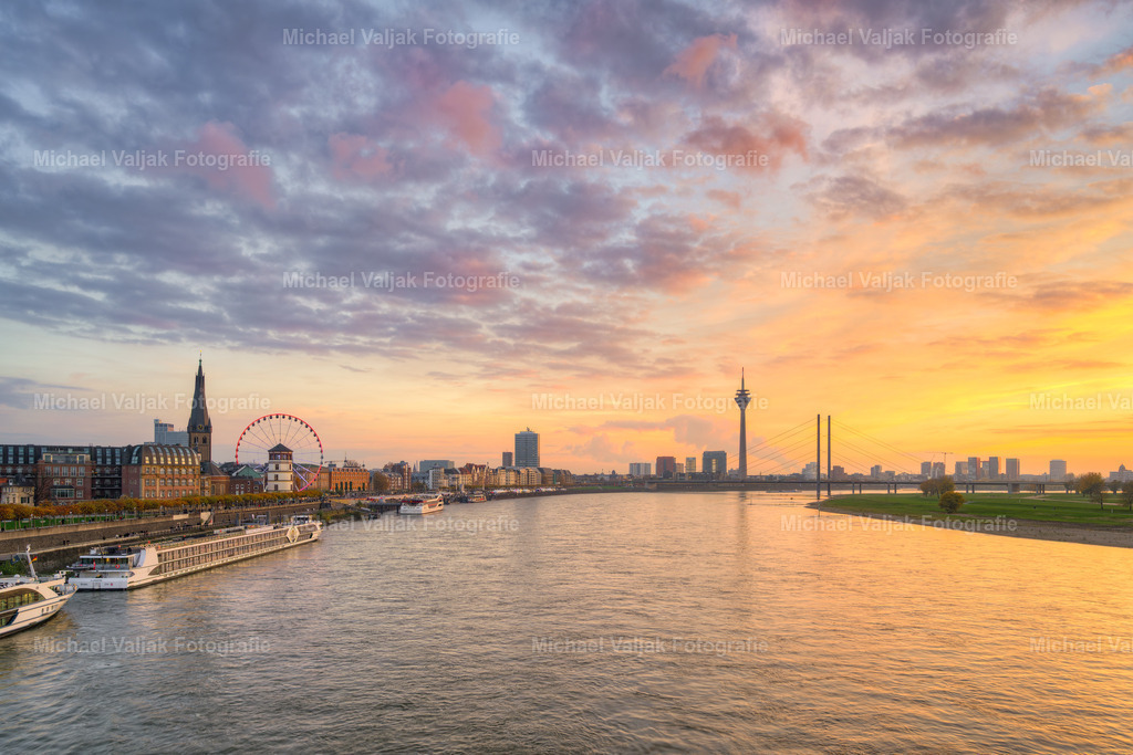 Die Düsseldorfer Skyline bei Sonnenuntergang | Von der Oberkasseler Brücke entfaltet sich im Abendlicht ein Panorama über Düsseldorf. Die Skyline erscheint im warmen Schimmer des Sonnenuntergangs, während am Burgplatz das Riesenrad seine Lichter entfaltet und einen lebendigen Akzent setzt. In weiter Ferne ist der Rheinturm zu erkennen, der die Silhouette der Stadt ergänzt und als markantes Wahrzeichen die Szene abrundet. So entsteht ein Bild, das die Vielfalt zwischen urbaner Moderne und städtischer Atmosphäre eindrucksvoll zeigt. - Realisiert mit Pictrs.com