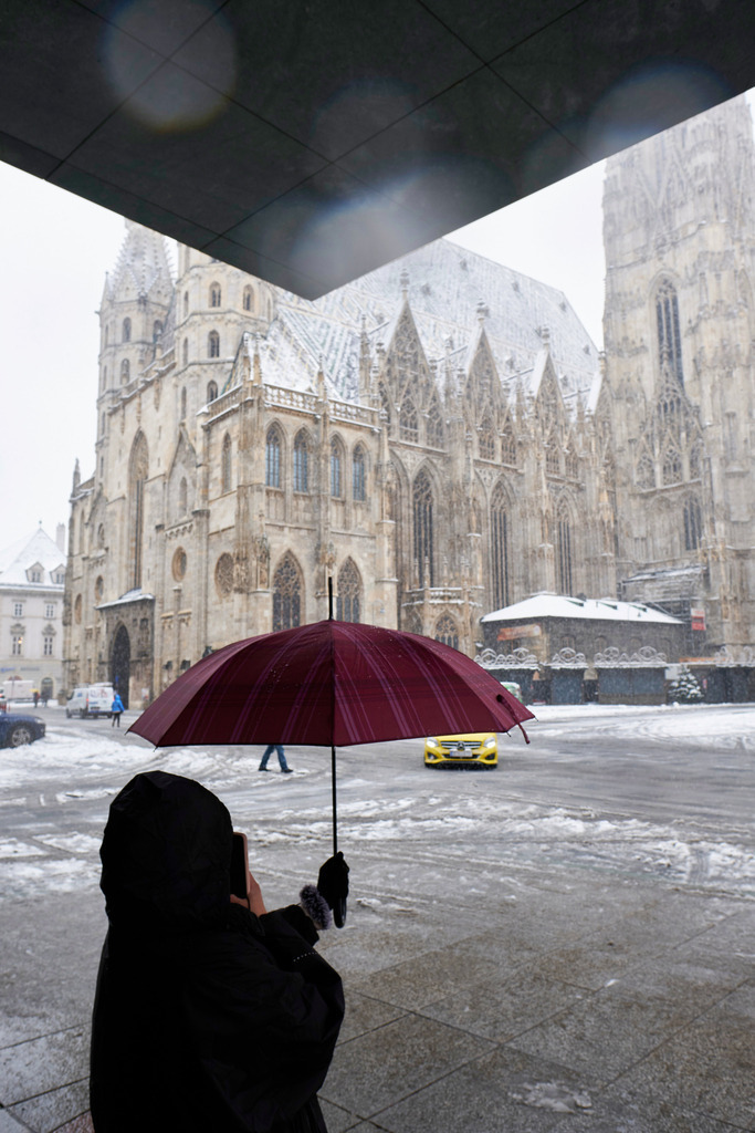 wartende Frau mit Schirm vor dem Stephansdom | Wien, Austria - December 09, 2021: Massnahmen zur Einschränkung von COVID-19; wartende Frau mit Schirm vor dem Stephansdom, im Hintergrund ein Taxi. - Realisiert mit Pictrs.com