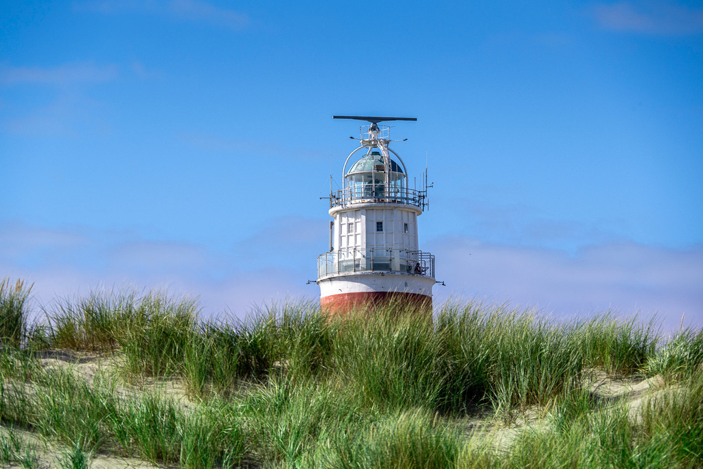 Detailansicht der Laterne des Leuchtturms Eierland hinter Dünen auf Texel | Nahaufnahme des historischen Leuchtturms Eierland an der Nordspitze der niederländischen Insel Texel. Die Vergrößerung zeigt die markante weiß-rote Turmspitze mit Galerie, Radar- und Lichtanlage, die stilvoll über den begrünten Dünengras-Kamm hervorragt. Die Aufnahme bei leicht bewölktem, blauem Himmel eignet sich perfekt für redaktionelle Beiträge über maritime Technik, Küstennavigation, Wahrzeichen, Inseltourismus an der Nordsee und den Urlaub in den Niederlanden. - Realisiert mit Pictrs.com