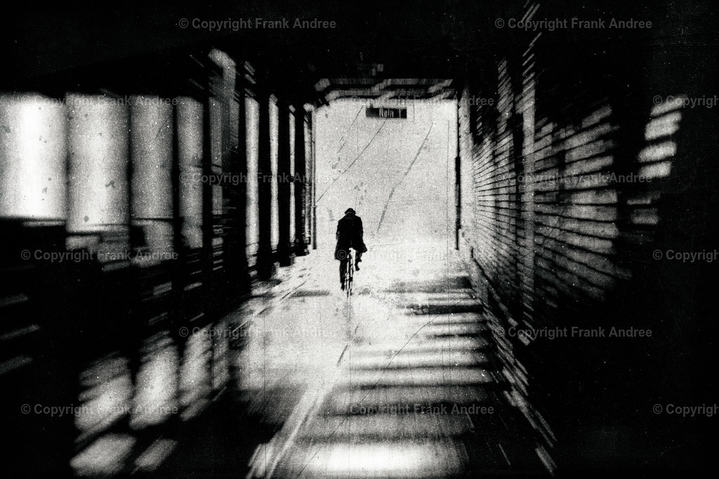 Radfahrer im Tunnel | Ein regnerischer Tag in Berlin. Die Straßen sind nass vom Regen. Ein Radfahrer mit Hut und Mantel fährt schnell durch einen Tunnel. Er fährt aus dem Tunnel hinaus ans Licht. Abstrakte schwarz-weiße Straßenfotografie aus Berlin. - Realisiert mit Pictrs.com