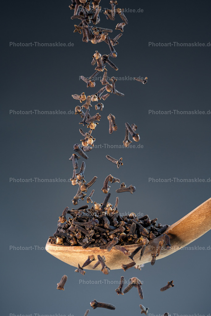 Gewürz in Bewegung - Tanz der Nelken | Gewürz in Bewegung - Tanz der Nelken. Kollektion: A spoon of tastefully seasoningsDas Bild zeigt getrocknete Gewürznelken, die in einer fließenden Bewegung von oben herabregnen und sich in einem elegant geschwungenen Holzlöffel sammeln. Die Farben sind warm und erdig – tiefes Dunkelbraun der Nelken, goldenes Holz und ein ruhiger, blaugrauer Hintergrund, der die Szene fast schwebend wirken lässt. Die Lichtführung ist weich, aber präzise, wodurch jede Nelke plastisch und detailreich hervorgehoben wird.Stil:Der Stil ist modern-minimalistisch mit einer Prise Food-Art-Fotografie. Die Komposition lebt von der Reduktion auf wenige Elemente, gepaart mit starker visueller Wirkung durch Licht, Bewegung und Struktur.Einsatzort:Dieses Bild würde sich ideal eignen für:Küchen und Essbereiche – als stilvolles Gewürzmotiv mit natürlicher Eleganz.Cafés oder Restaurants – besonders in Bereichen mit Gewürz- oder Naturthema.Gewürzläden oder Feinkostgeschäfte – als dekorativer Eyecatcher.Moderne Wohnräume – z.B. in Kombination mit weiteren Naturfotografien in einer Galerie-Wand. - Realisiert mit Pictrs.com