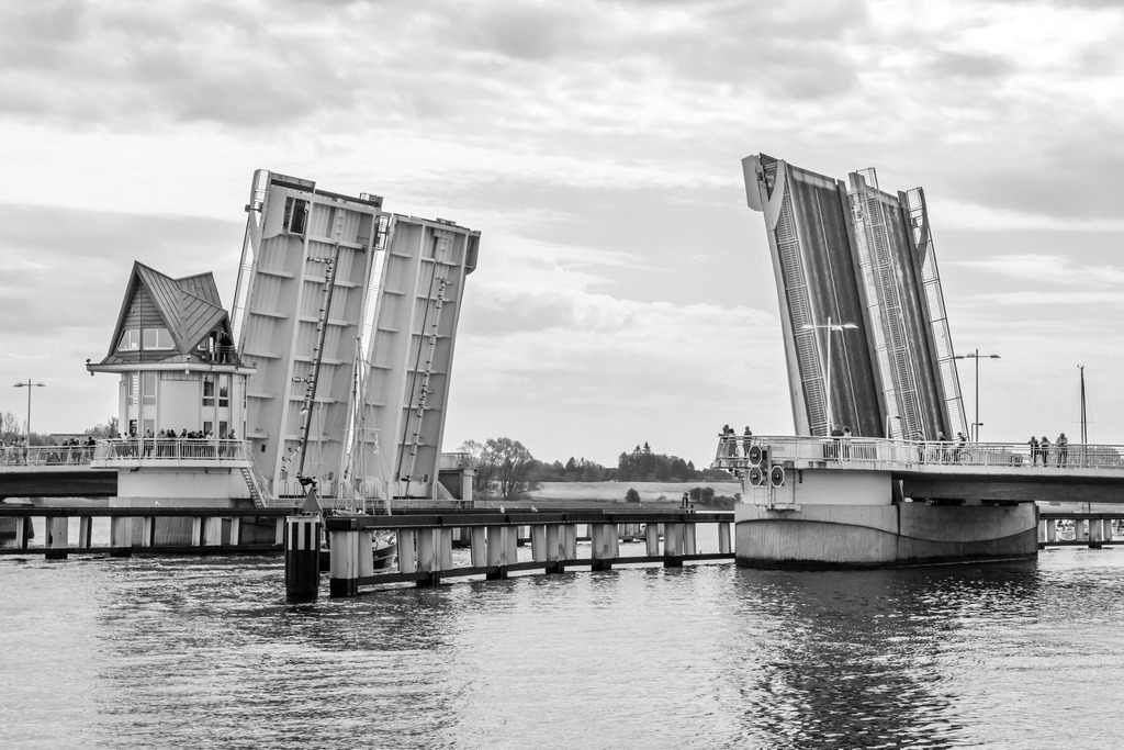 Wandbild: Geöffnete Schleibrücke in Kappeln in Schwarz-Weiß | Erleben Sie die technische Raffinesse und das maritime Flair von Kappeln mit dieser beeindruckenden Schwarz-Weiß-Aufnahme der geöffneten Schleibrücke. Das Bild zeigt die Brücke in gehobenem Zustand, um Segelbooten die Durchfahrt zu ermöglichen. Menschen sind auf der Brücke und in der Nähe des Steuerhauses zu sehen, was die Lebendigkeit der Szene unterstreicht.Dieses Kunstwerk, erhältlich auf Leinwand, Alu-Dibond, Acrylglas, Fineart und als Akustikbild, bringt die beeindruckende Architektur und Funktionalität der Schleibrücke direkt in Ihr Zuhause. - Realisiert mit Pictrs.com