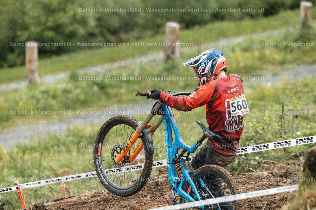 Mountainbiker im Abfahrtssprint | Ein Mountainbiker in voller Ausrüstung rast einen steilen, verschneiten Hang hinunter. Er trägt einen roten Trikot mit der Nummer 560 und einen blauen Helm. Das Gelände ist mit Gras und vereinzelten Büschen bedeckt. Im Hintergrund sind Bäume und ein verschneiter Gipfel zu sehen. - Realisiert mit Pictrs.com