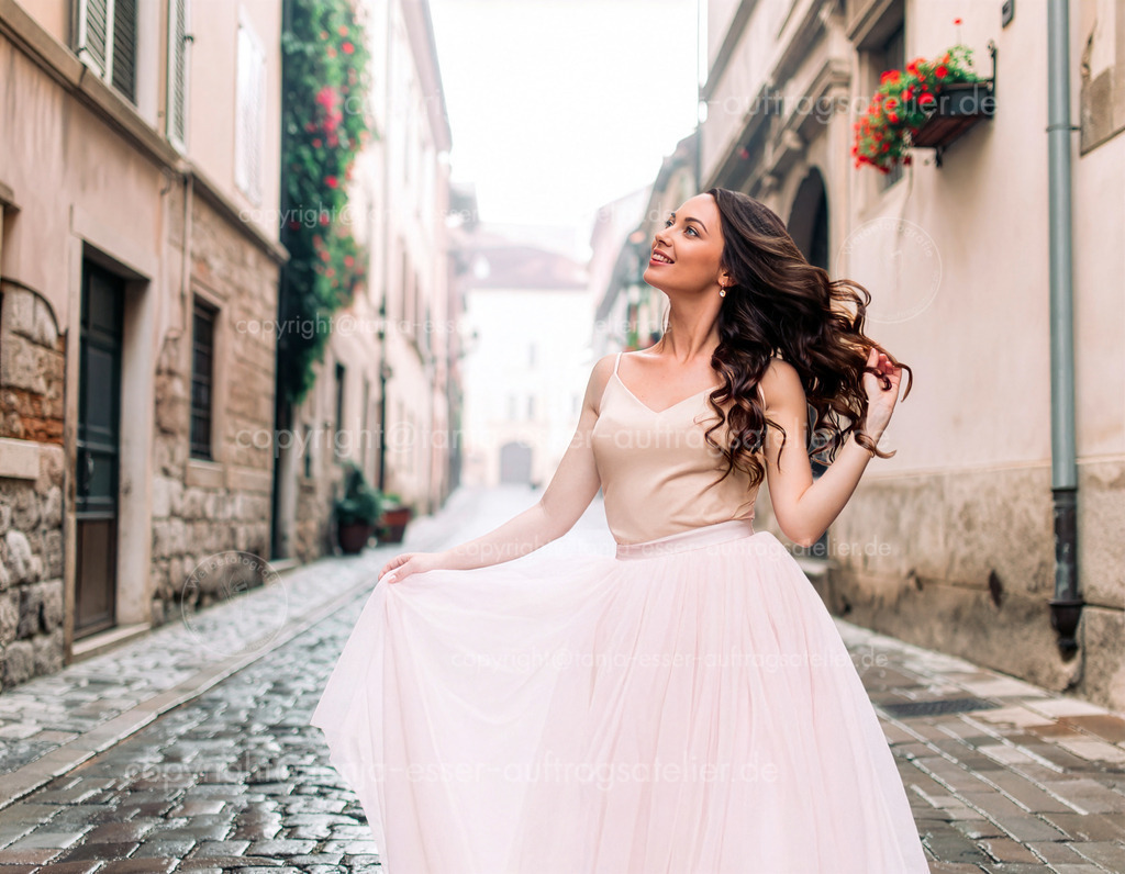 Verspielte schöne Frau mit langen dunklen Haaren tanzt in der Stadt | Eine schöne Frau mit langen, dunkel gewellten Haaren posiert voller Lebensfreude in einer Altstadtgasse. Sie trägt einen hellrosa Tüllrock. Romantisch, feminin, verspielt und verträumt. Ausgearbeitetes Bild einer generativen KI.