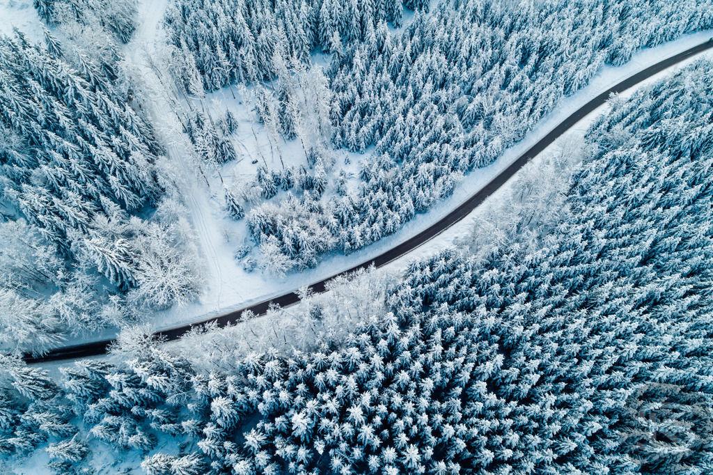 Winterwald aus der Luft – Verschneite Straße durch gefrorenen Nadelwald | Diese Drohnenaufnahme zeigt eine kurvenreiche Straße, die sich durch einen dicht verschneiten Nadelwald schlängelt. Aus der Vogelperspektive wird die geometrische Struktur sichtbar: Die organische Linie der Straße bildet einen starken Kontrast zu den vertikalen Mustern der schneebedeckten Bäume. Die kalten Blautöne verstärken die winterliche Atmosphäre und verleihen dem Wald eine fast abstrakte Qualität. - Realisiert mit Pictrs.com